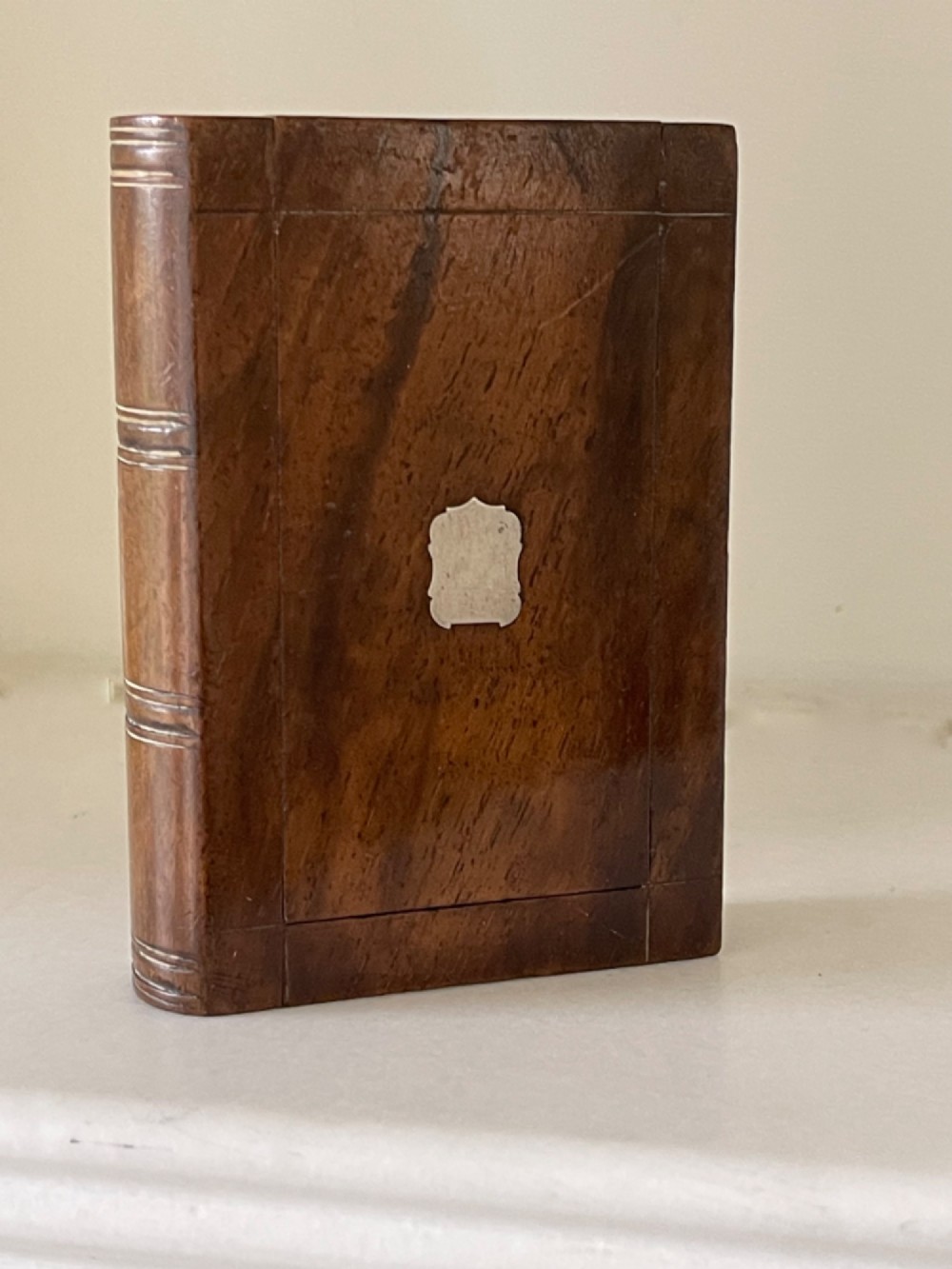 A small warm brown wooden box shaped to resemble a bound book with a silver shield cartouche inlaid on the front lid.