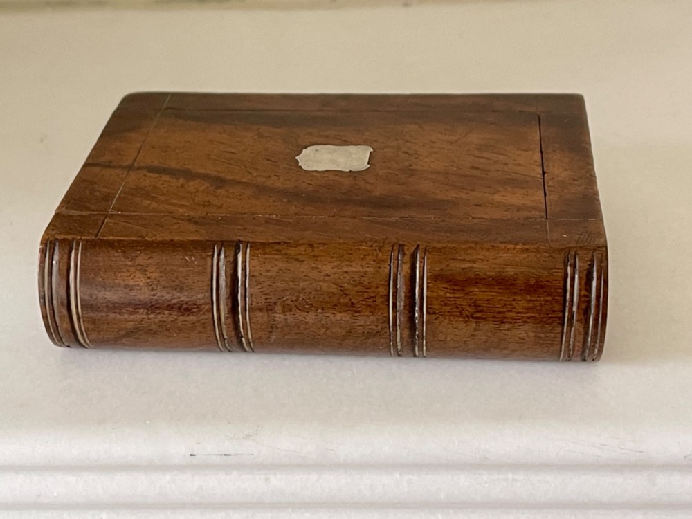 A small warm brown wooden box shaped to resemble a bound book with a silver shield cartouche inlaid on the front lid. View II