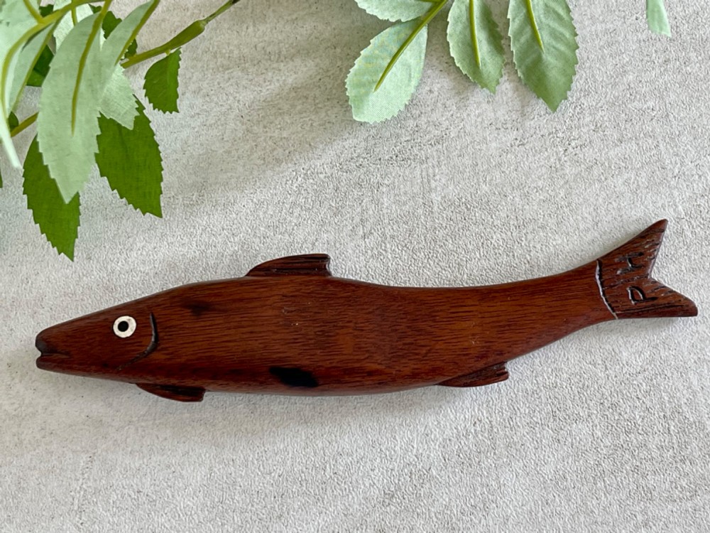 A hand-carved dark wood knitting sheath shaped like a fish, featuring an inlaid eye and carved initials near the tail fin.