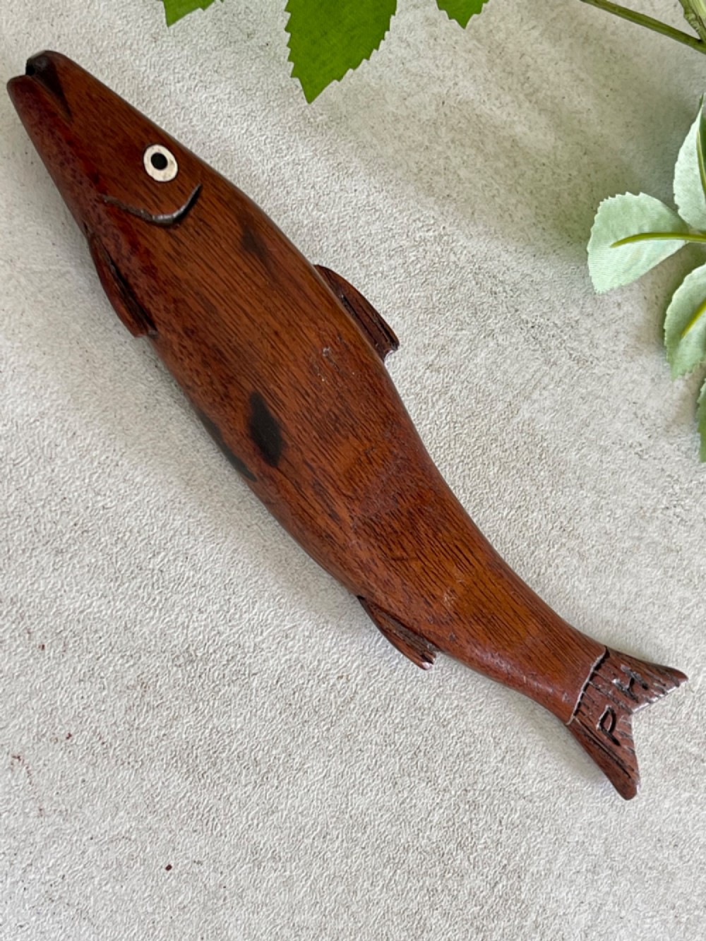 A hand-carved dark wood knitting sheath shaped like a fish, featuring an inlaid eye and carved initials near the tail fin. View V
