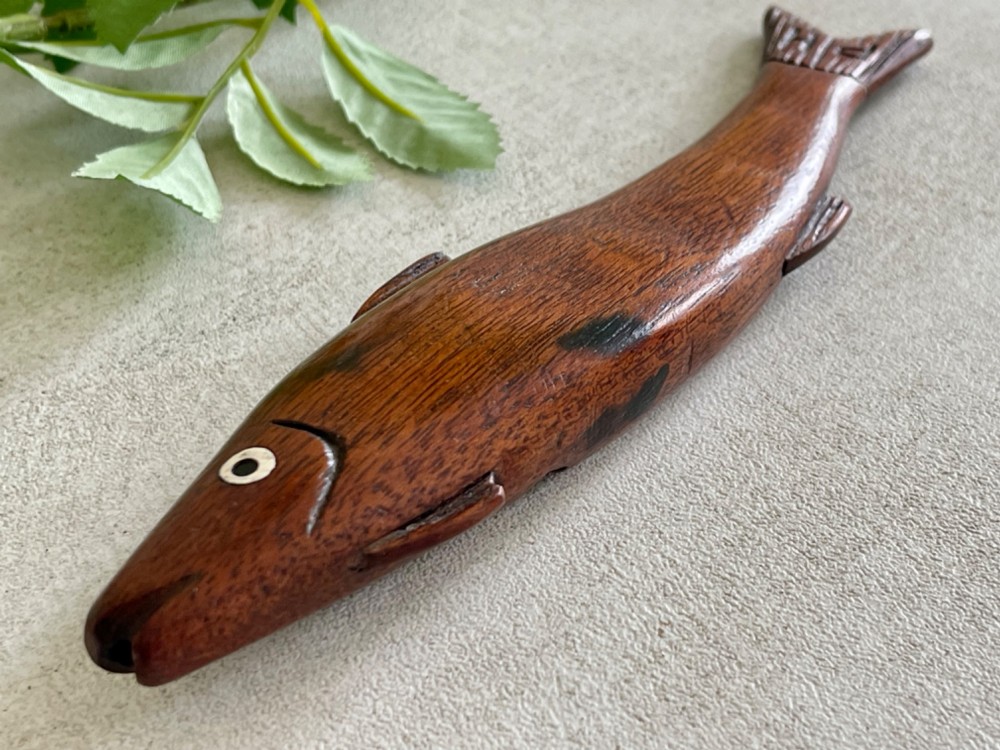 A hand-carved dark wood knitting sheath shaped like a fish, featuring an inlaid eye and carved initials near the tail fin. View VII