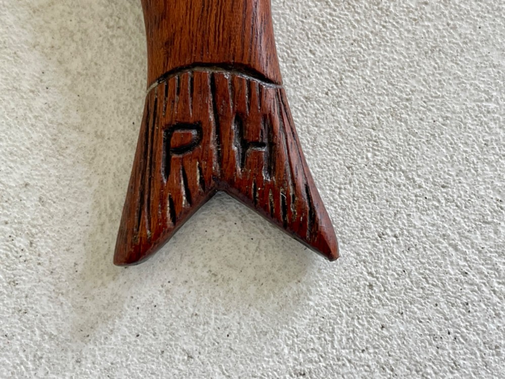 A hand-carved dark wood knitting sheath shaped like a fish, featuring an inlaid eye and carved initials near the tail fin. View VIII