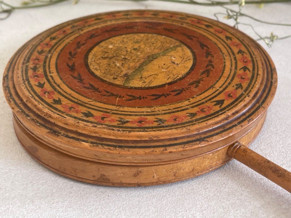 A circular wooden travelling mirror featuring concentric red and black decorative bands around the lid and a turned wood handle. View V