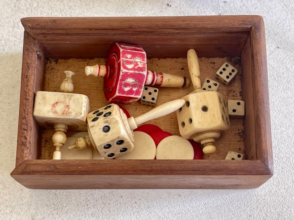 A rectangular wooden box holding hand-carved bone teetotum spinning tops, miniature dice, and red and white counter discs.