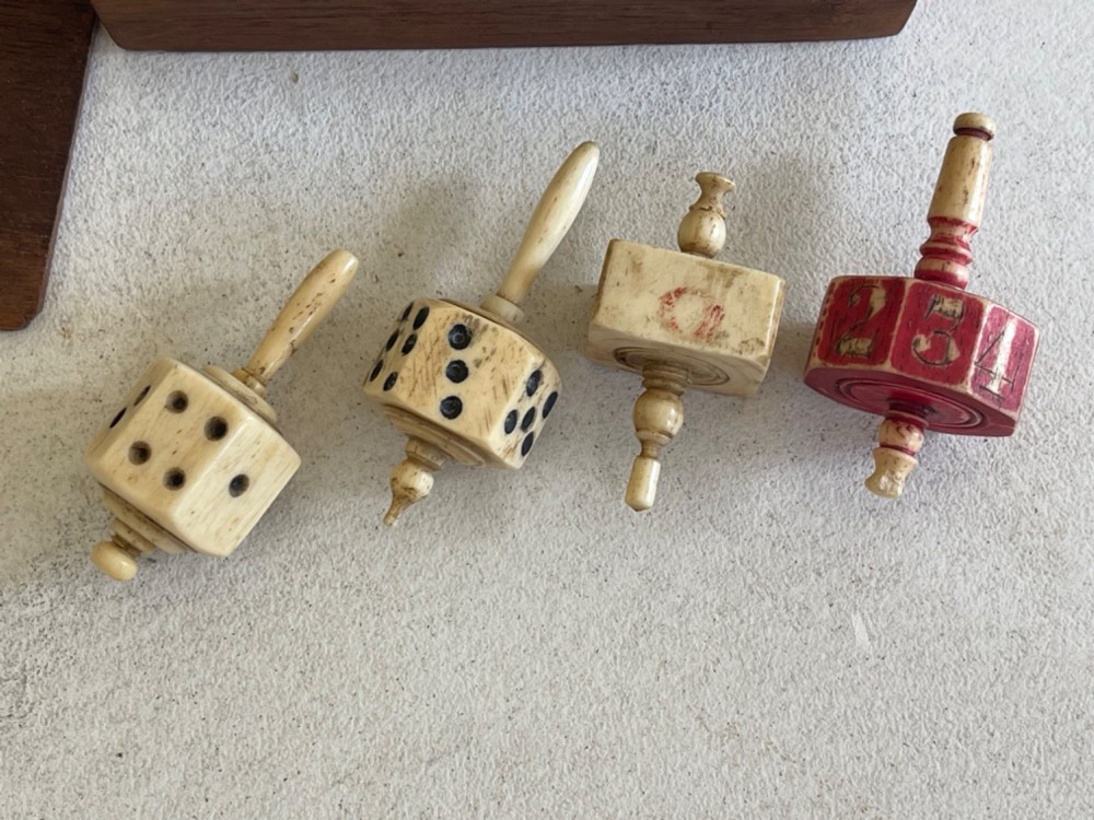 A rectangular wooden box holding hand-carved bone teetotum spinning tops, miniature dice, and red and white counter discs. View II