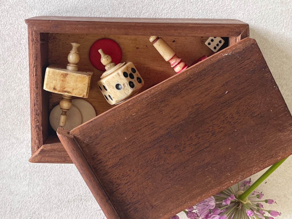 A rectangular wooden box holding hand-carved bone teetotum spinning tops, miniature dice, and red and white counter discs. View III