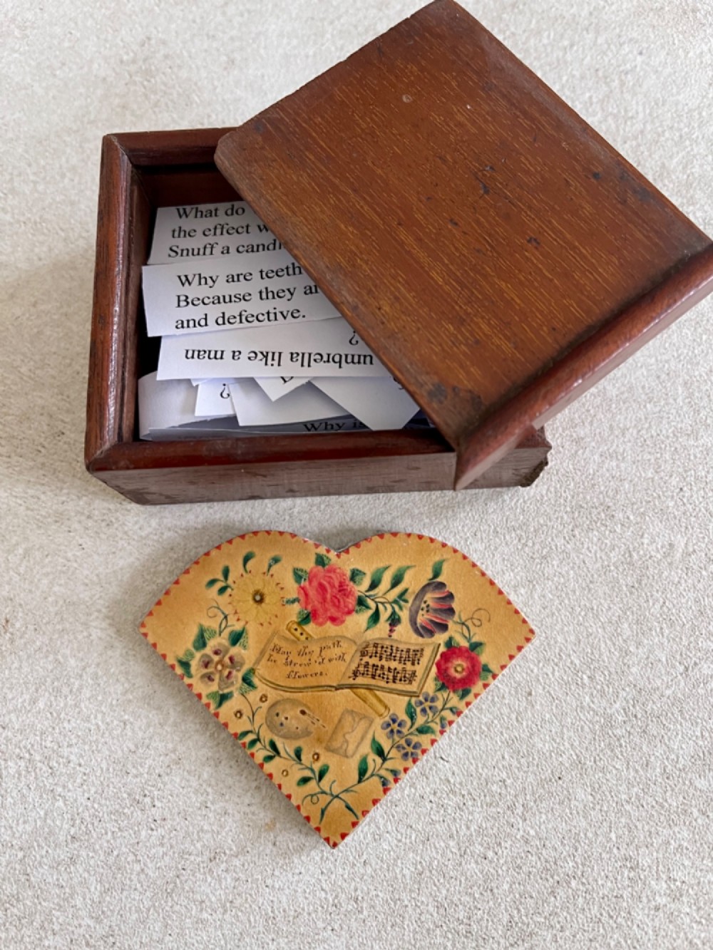 An open small wooden box containing printed paper riddle slips set behind a heart-shaped printed card with floral motifs.