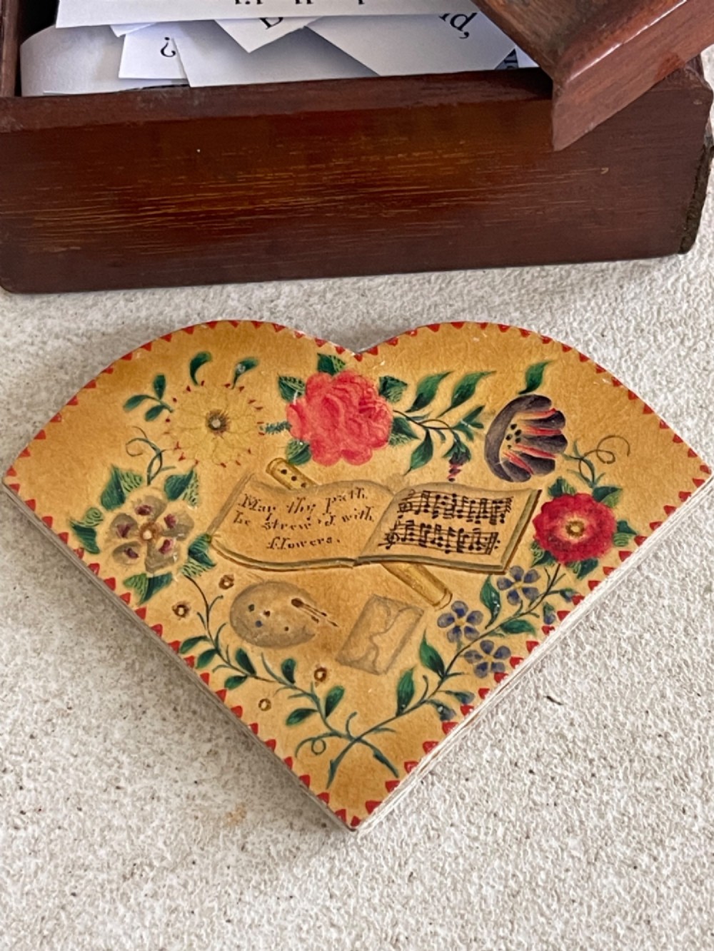 An open small wooden box containing printed paper riddle slips set behind a heart-shaped printed card with floral motifs. View III