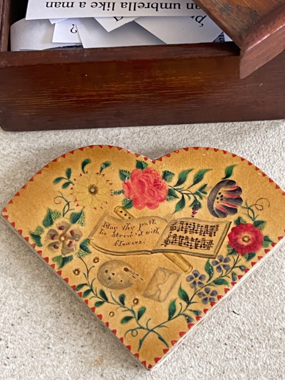An open small wooden box containing printed paper riddle slips set behind a heart-shaped printed card with floral motifs. View VI