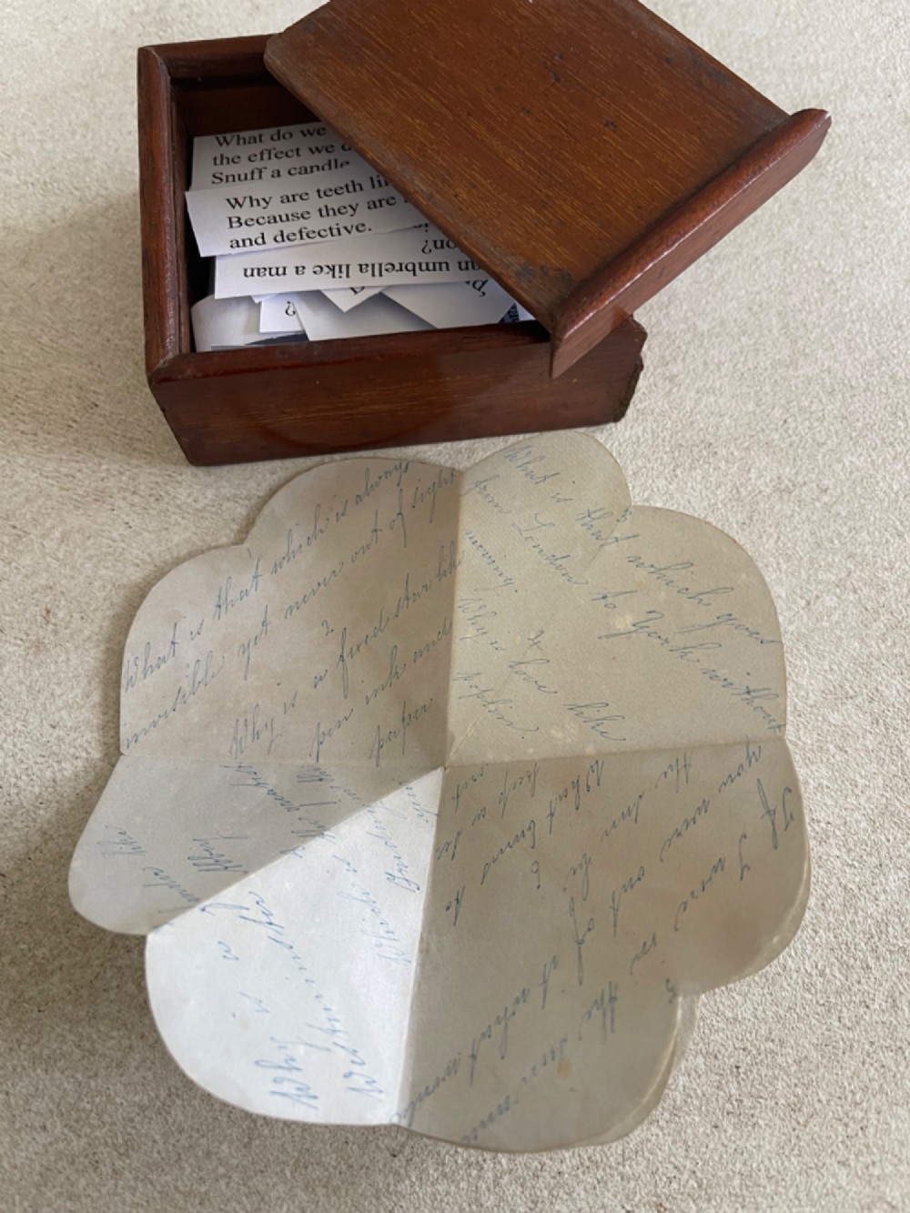 An open small wooden box containing printed paper riddle slips set behind a heart-shaped printed card with floral motifs. View VIII