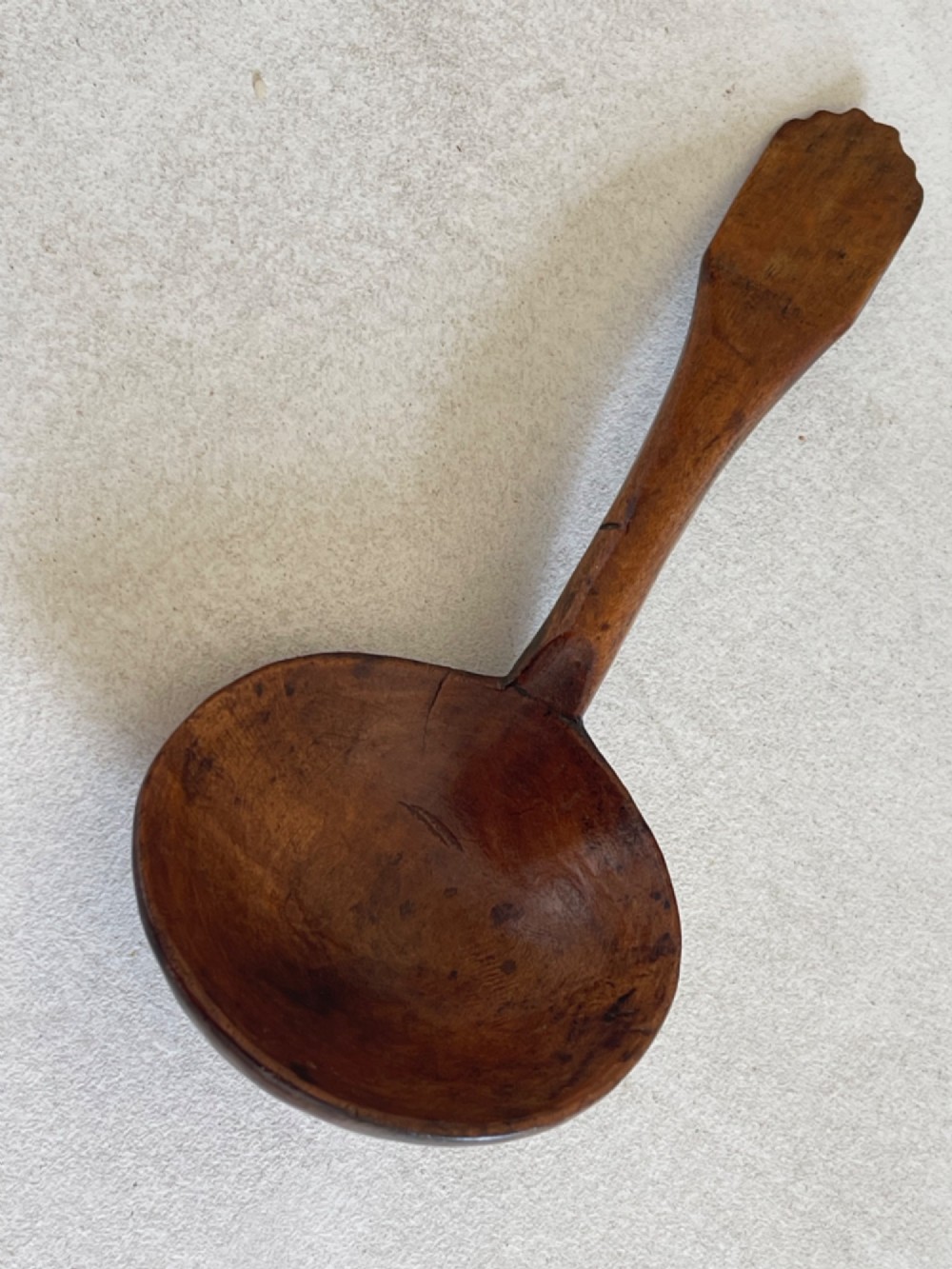A hand-carved antique treen wooden ladle featuring a deep circular bowl and a shaped handle with decorative notched detailing. View V
