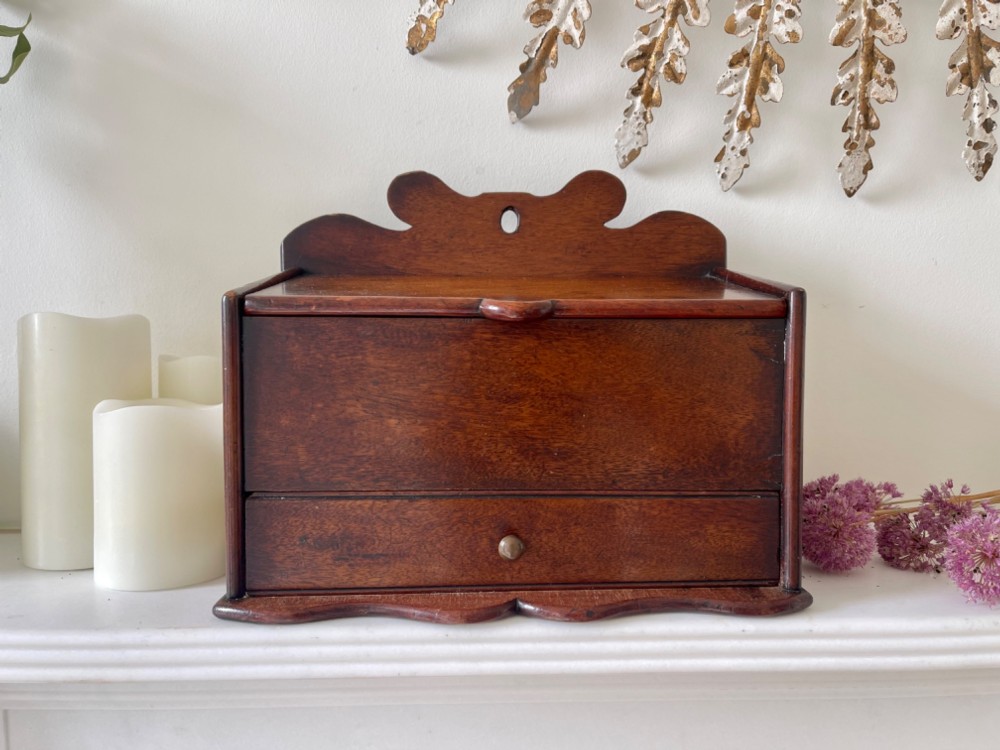 An antique dark mahogany wooden candle box featuring a shaped backboard with a hanging hole, a hinged top lid, a lower drawer with a metal knob, and a shaped apron base.