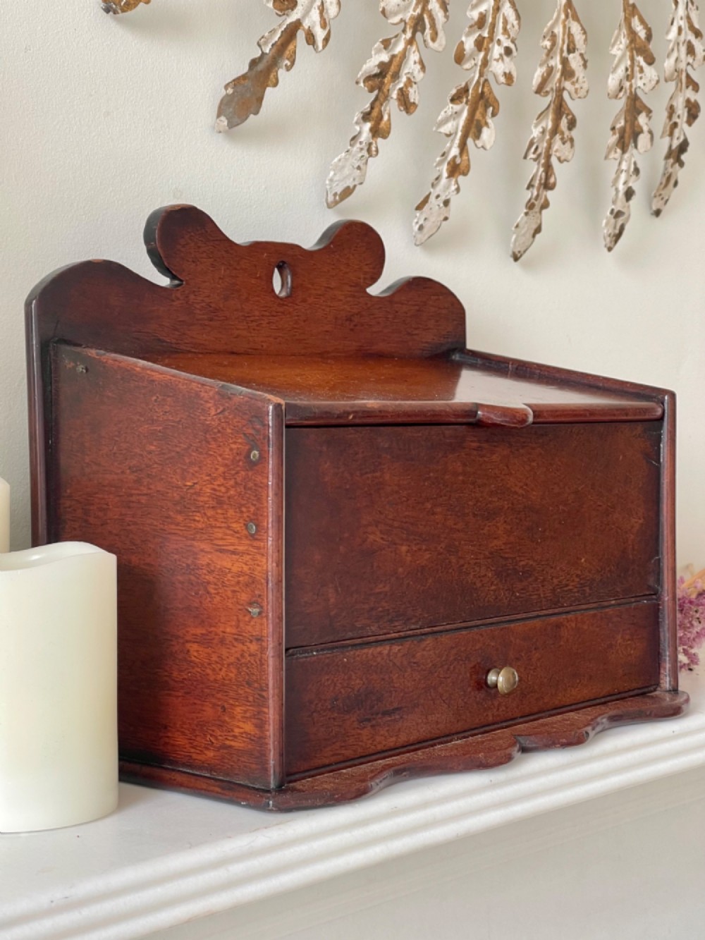 An antique dark mahogany wooden candle box featuring a shaped backboard with a hanging hole, a hinged top lid, a lower drawer with a metal knob, and a shaped apron base. View II