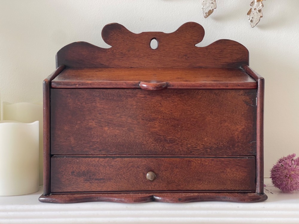 An antique dark mahogany wooden candle box featuring a shaped backboard with a hanging hole, a hinged top lid, a lower drawer with a metal knob, and a shaped apron base. View IV
