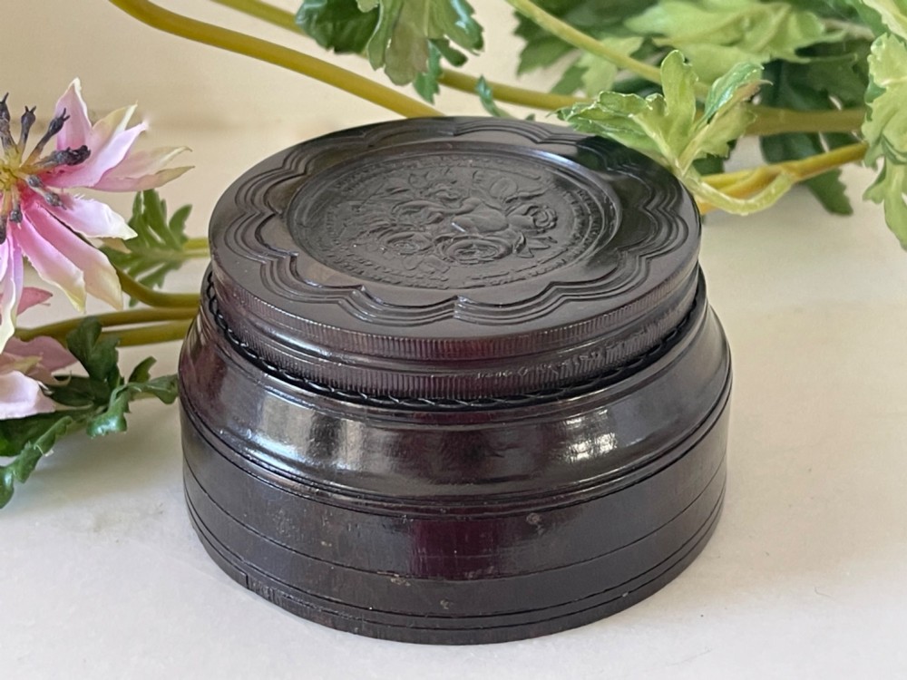 A circular dark brown engine-turned box featuring a central raised floral relief medallion on the lid.