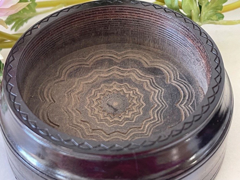 A circular dark brown engine-turned box featuring a central raised floral relief medallion on the lid. View V
