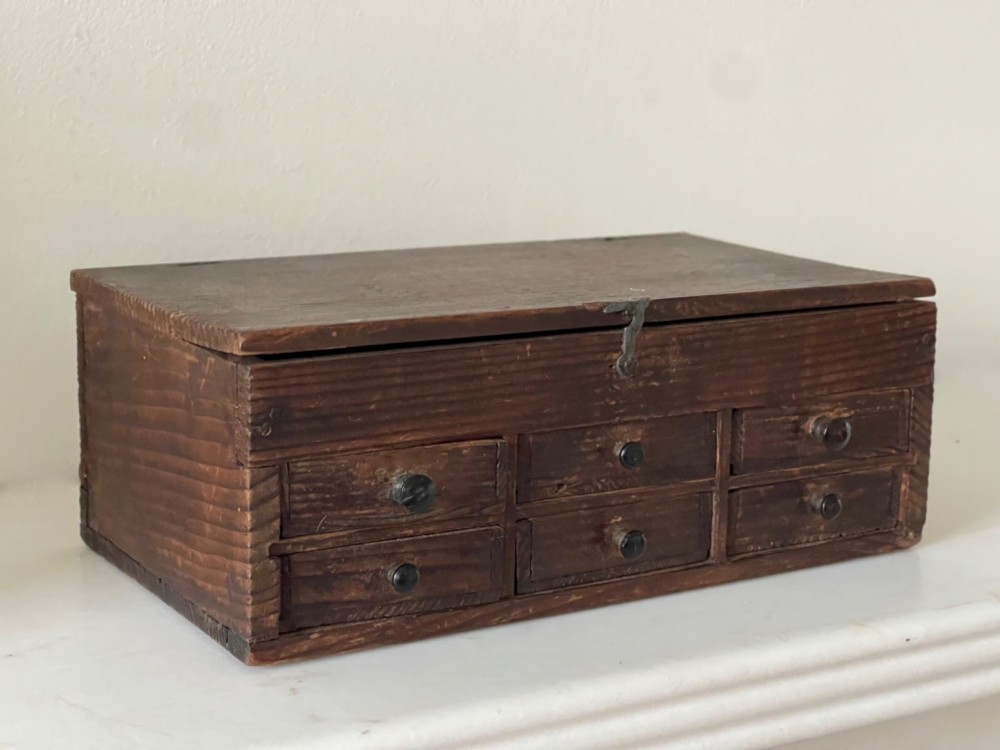 A dark pine antique watchmakers box featuring a hinged top lid, metal clasp, and six small wooden drawers with turned knobs.