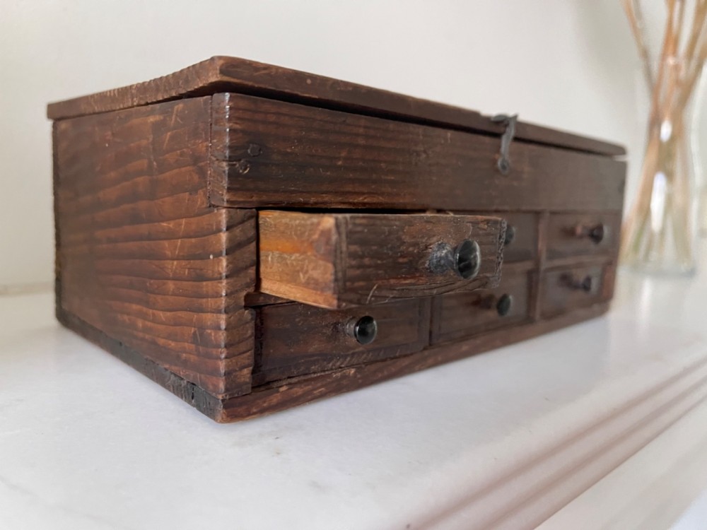 A dark pine antique watchmakers box featuring a hinged top lid, metal clasp, and six small wooden drawers with turned knobs. View II