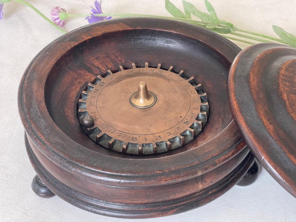 A dark turned wood circular tabletop roulette game featuring a stepped fitted lid with a turned central finial raised on bun feet. View II