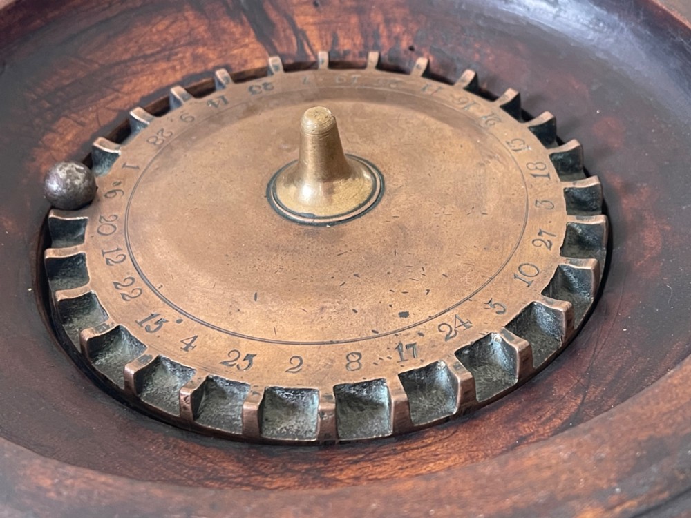 A dark turned wood circular tabletop roulette game featuring a stepped fitted lid with a turned central finial raised on bun feet. View V