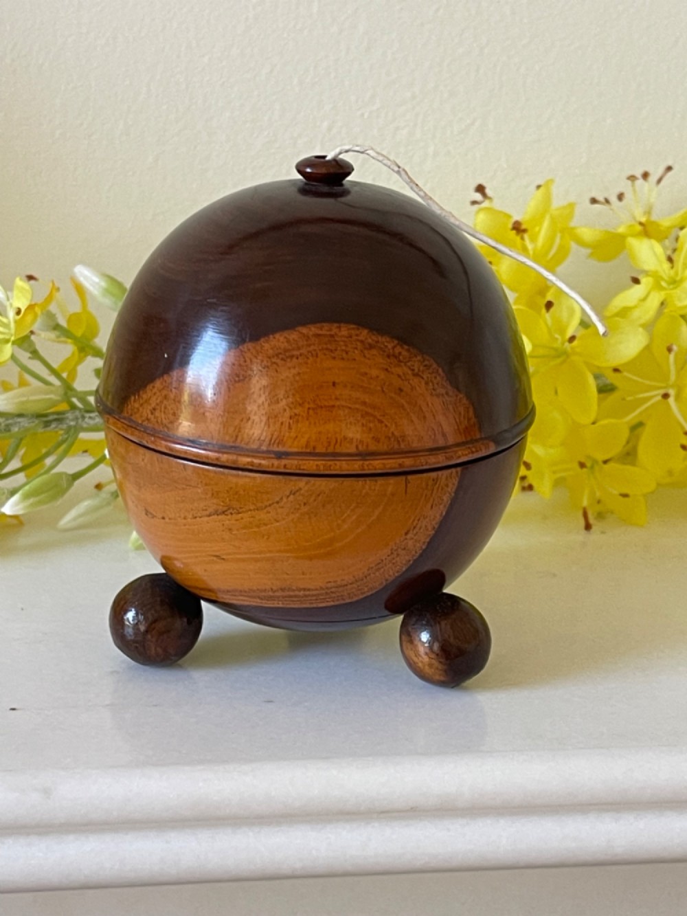 A turned dark wood spherical string box resting on three small ball feet with a thread emerging from the top aperture.