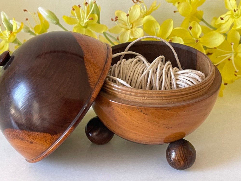 A turned dark wood spherical string box resting on three small ball feet with a thread emerging from the top aperture. View II