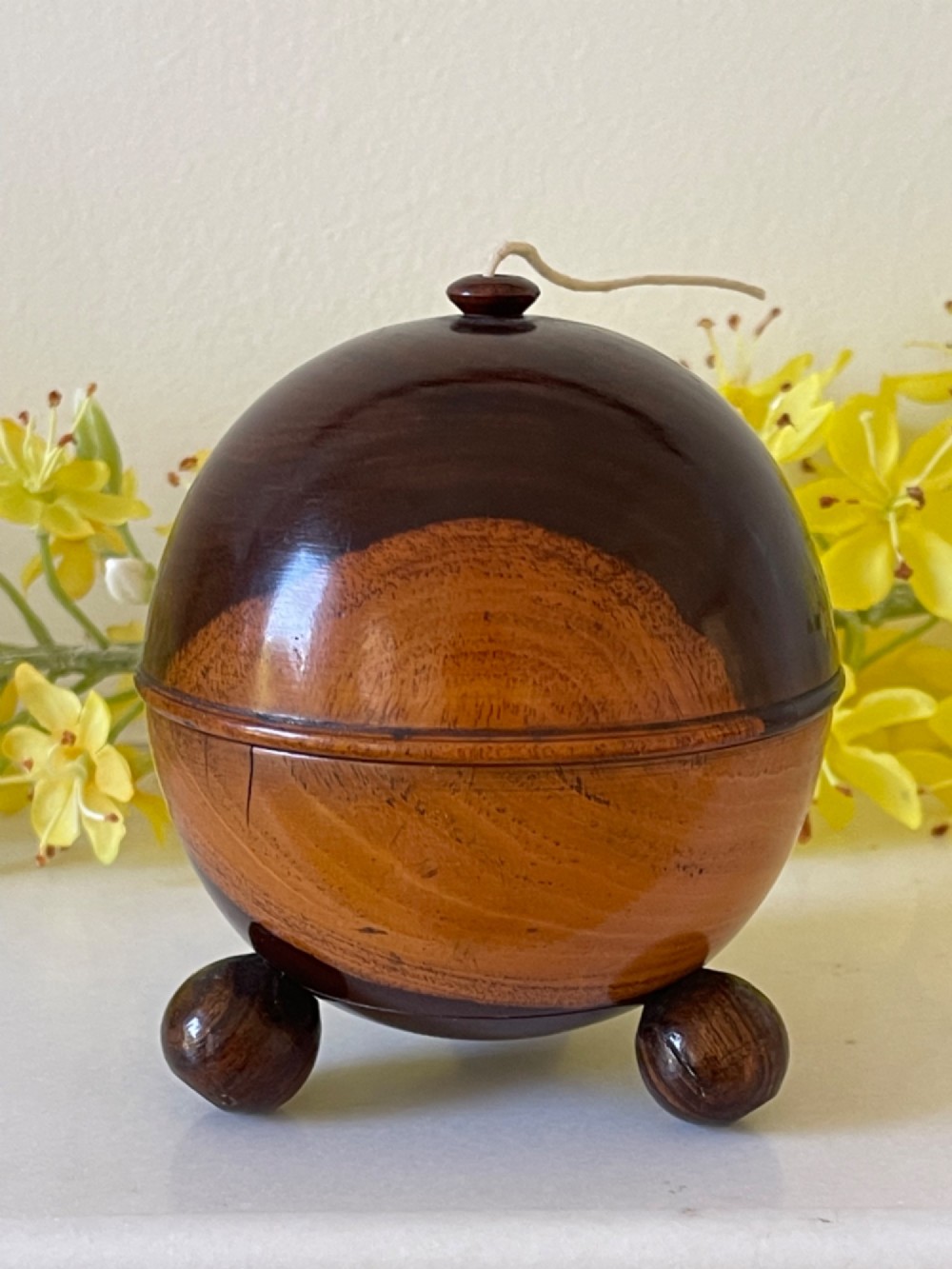 A turned dark wood spherical string box resting on three small ball feet with a thread emerging from the top aperture. View III
