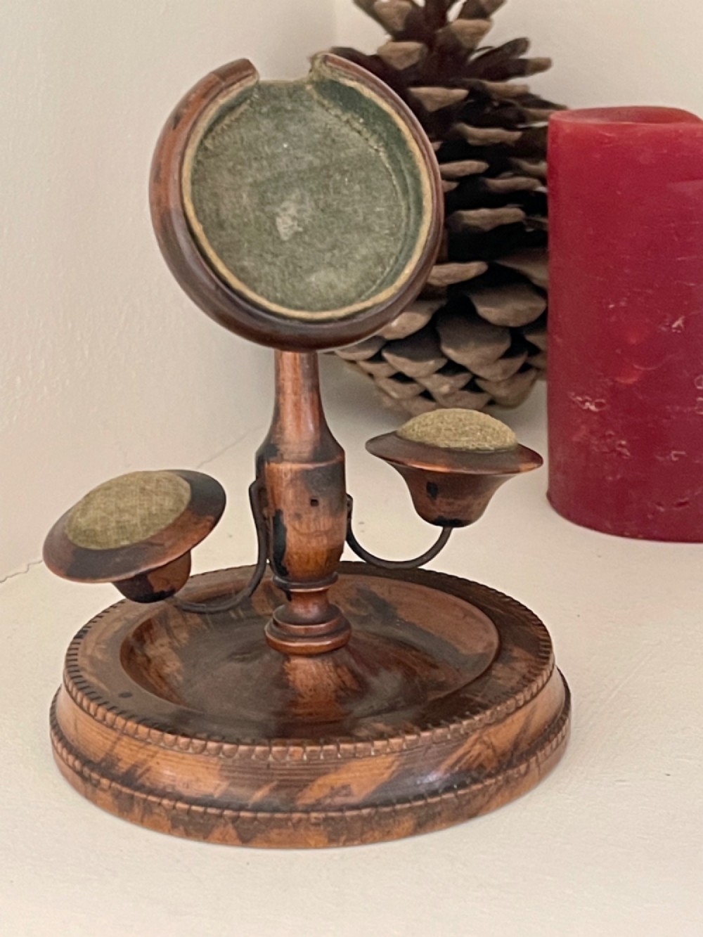 An antique turned wood pocket watch stand with a green velvet lined circular holder and two integrated side pincushions on a dished base.