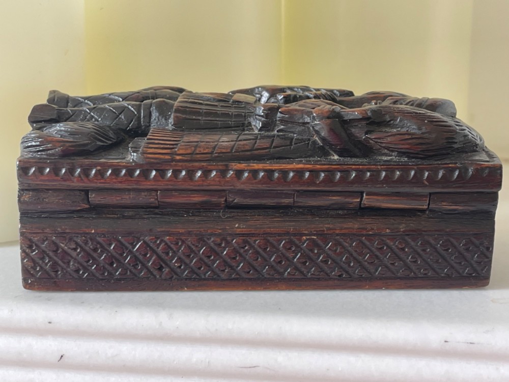 A dark carved wooden table snuff box lid depicting a Scottish Highlander in a kilt carrying a stag across his shoulders with a hound at his feet. View II