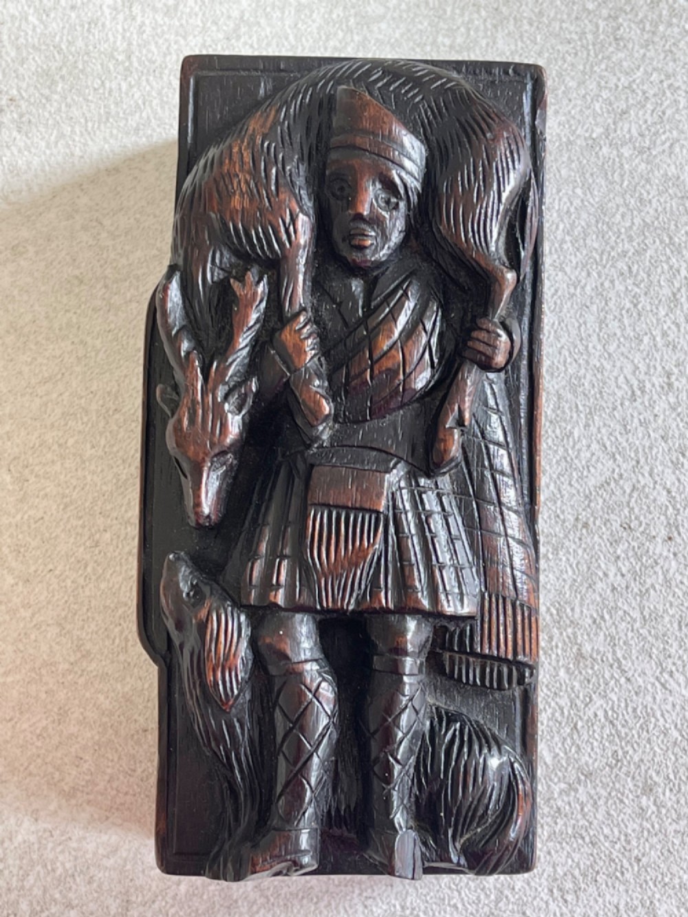 A dark carved wooden table snuff box lid depicting a Scottish Highlander in a kilt carrying a stag across his shoulders with a hound at his feet. View IV