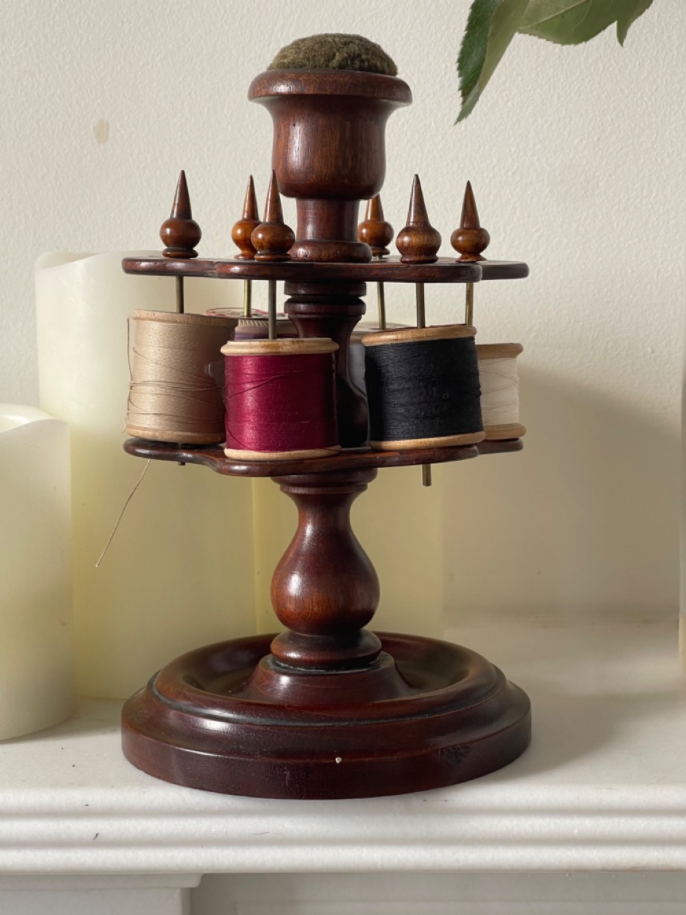 A turned dark mahogany cotton reel stand featuring a central pillar topped with a green fabric pin cushion and surrounded by brass spindles holding wooden thread spools.