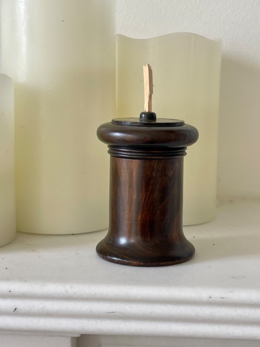 A small turned dark wooden go-to-bed match holder featuring a cylindrical body, a moulded cap, and a match inserted in the centre top.