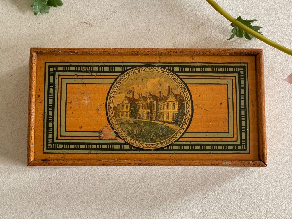 A rectangular wooden box lid featuring a central circular printed vignette of a country house surrounded by geometric green and black borders on an ochre surface.