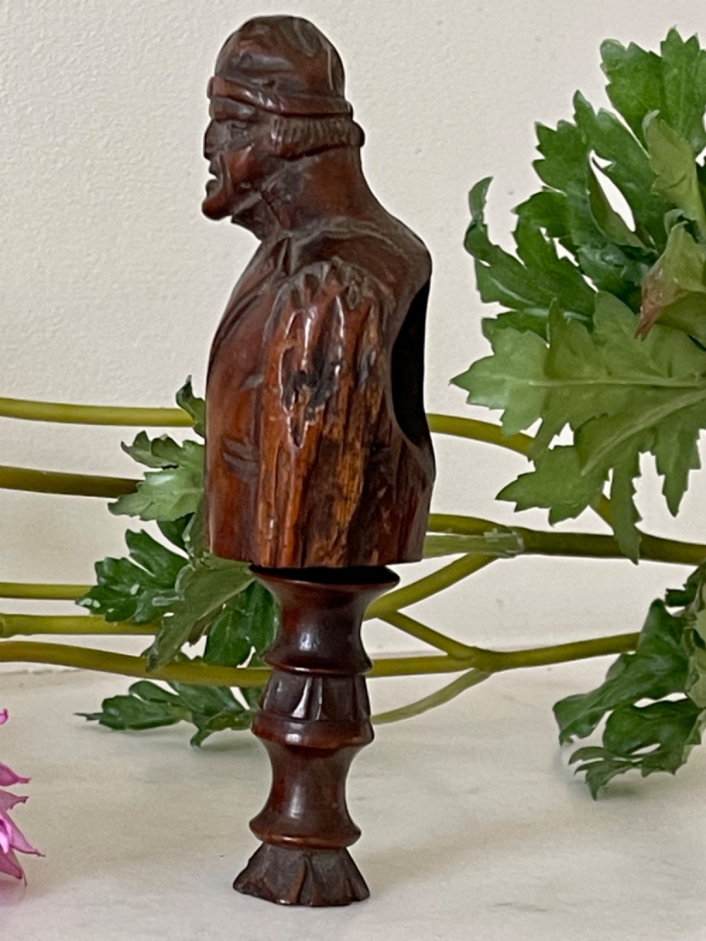 A dark carved wooden treen nutcracker featuring a male bust wearing a cap and jacket atop a turned pedestal handle. View II