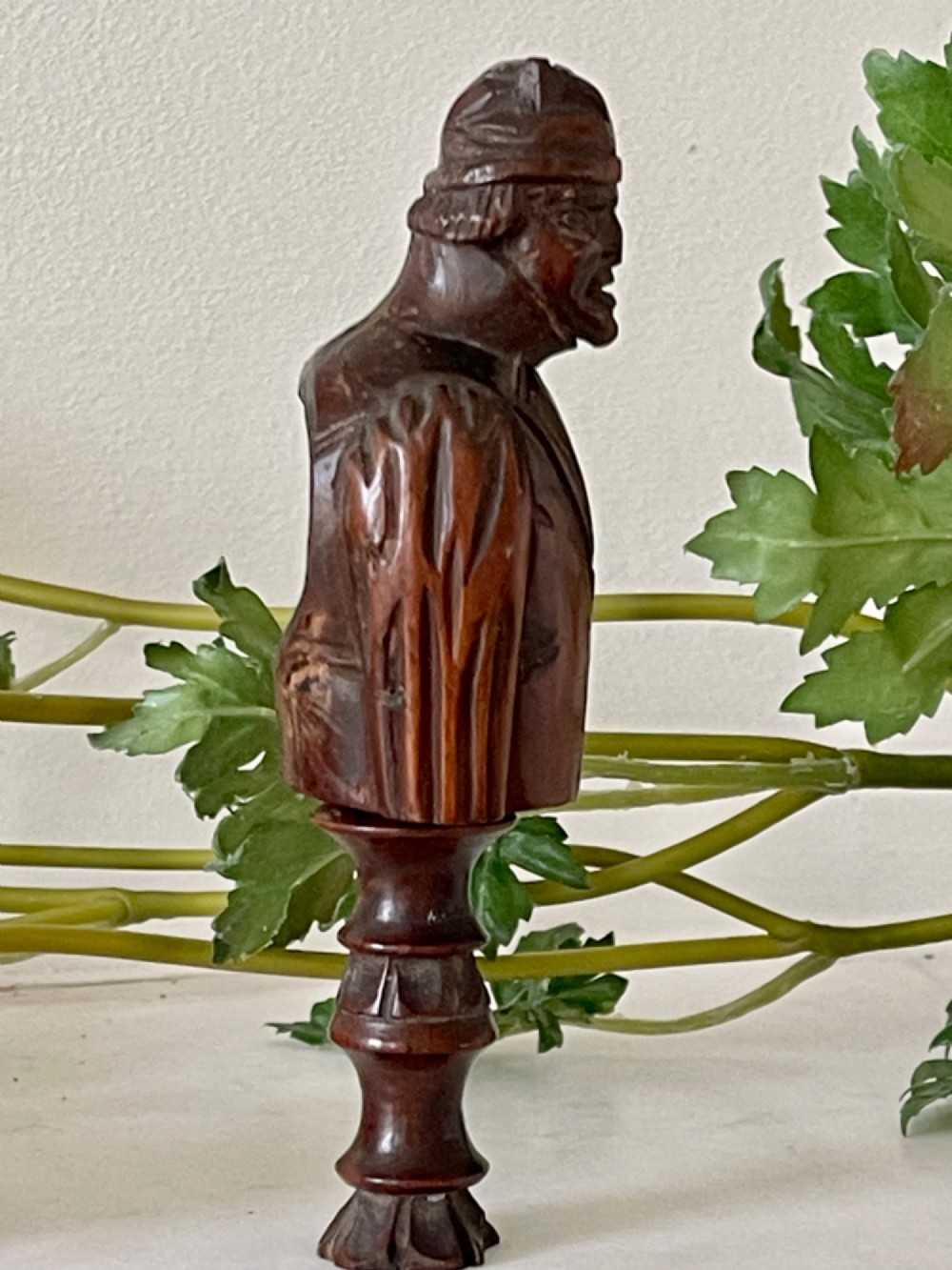 A dark carved wooden treen nutcracker featuring a male bust wearing a cap and jacket atop a turned pedestal handle. View V