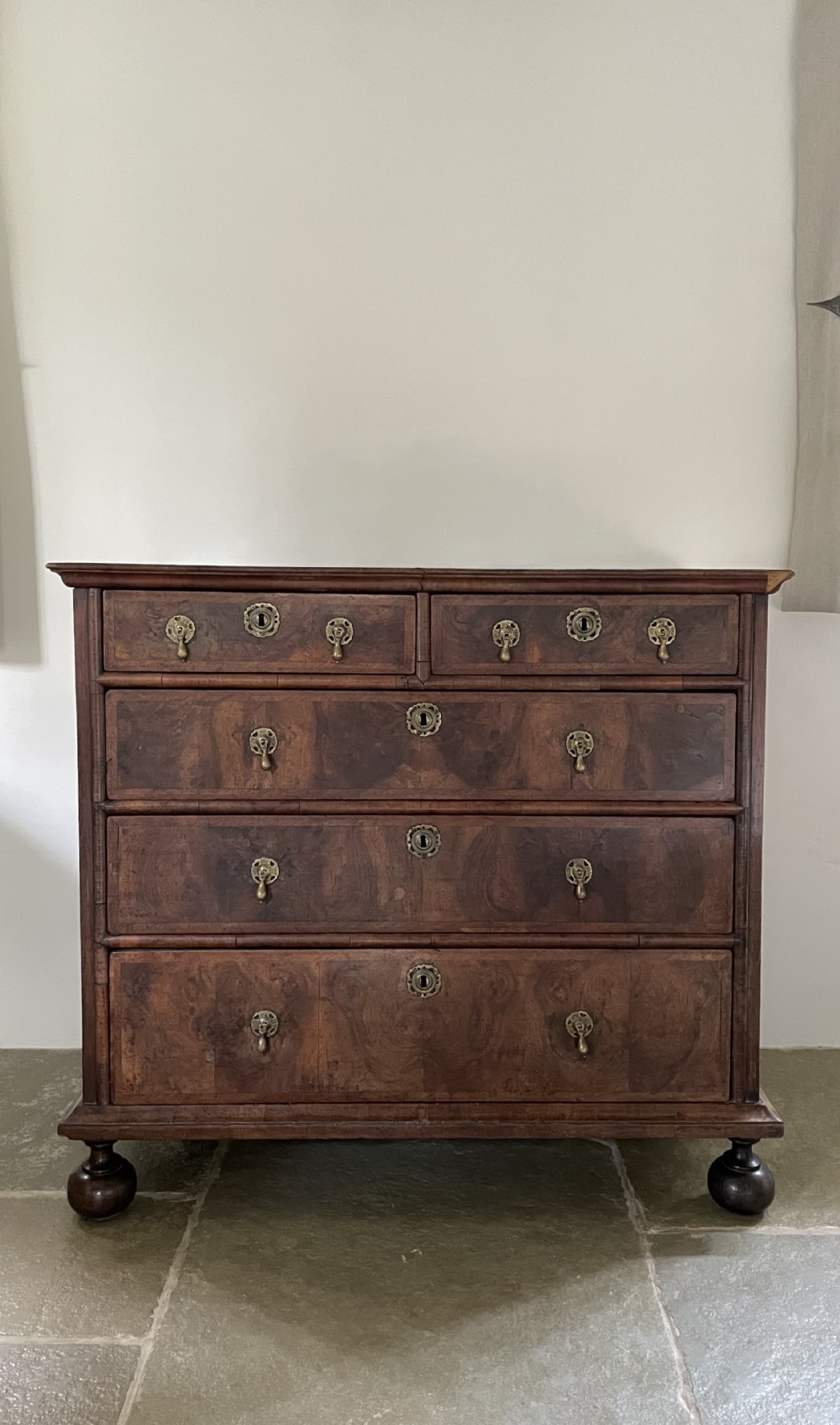A late 17th-century walnut chest of drawers featuring brass teardrop handles, keyhole escutcheons, crossbanded veneer drawer fronts, and turned bun feet.