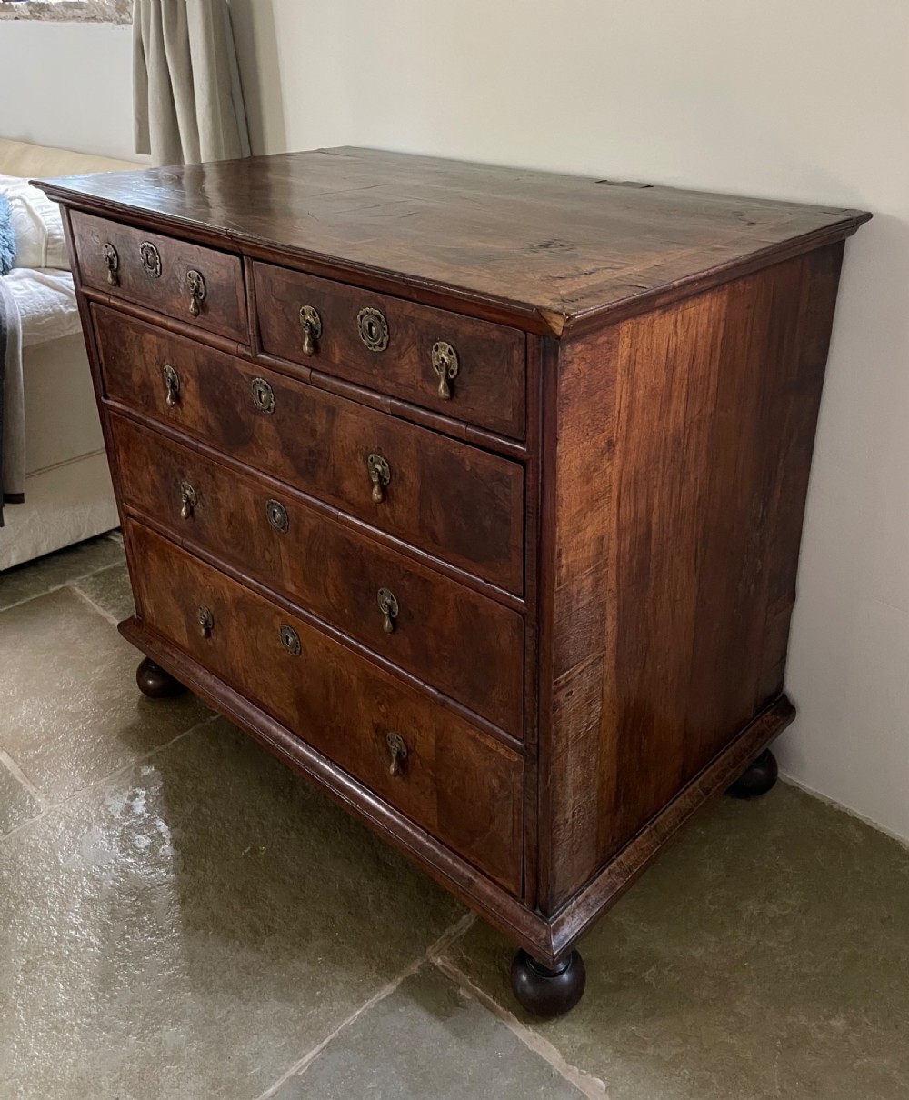A late 17th-century walnut chest of drawers featuring brass teardrop handles, keyhole escutcheons, crossbanded veneer drawer fronts, and turned bun feet. View V