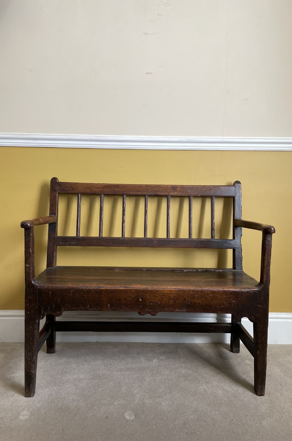 A dark oak Queen Anne period bench featuring open armrests, a vertical spindle backrest, a wooden plank seat, and a shaped apron.