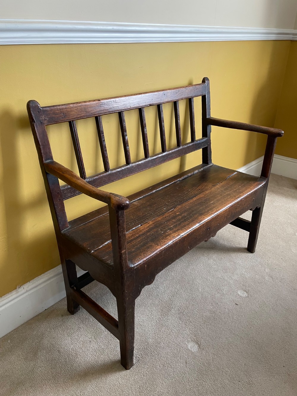 A dark oak Queen Anne period bench featuring open armrests, a vertical spindle backrest, a wooden plank seat, and a shaped apron. View II