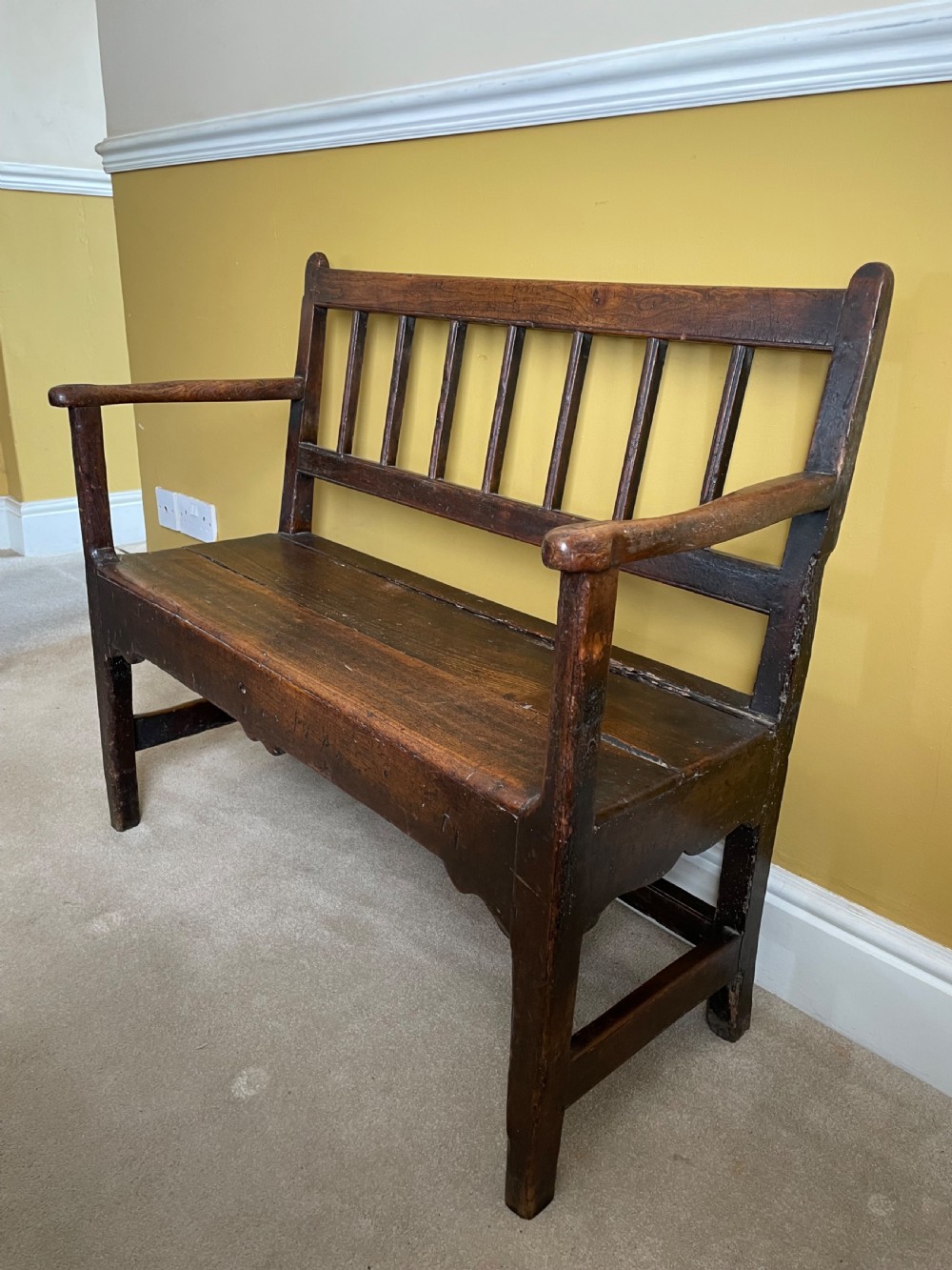 A dark oak Queen Anne period bench featuring open armrests, a vertical spindle backrest, a wooden plank seat, and a shaped apron. View III