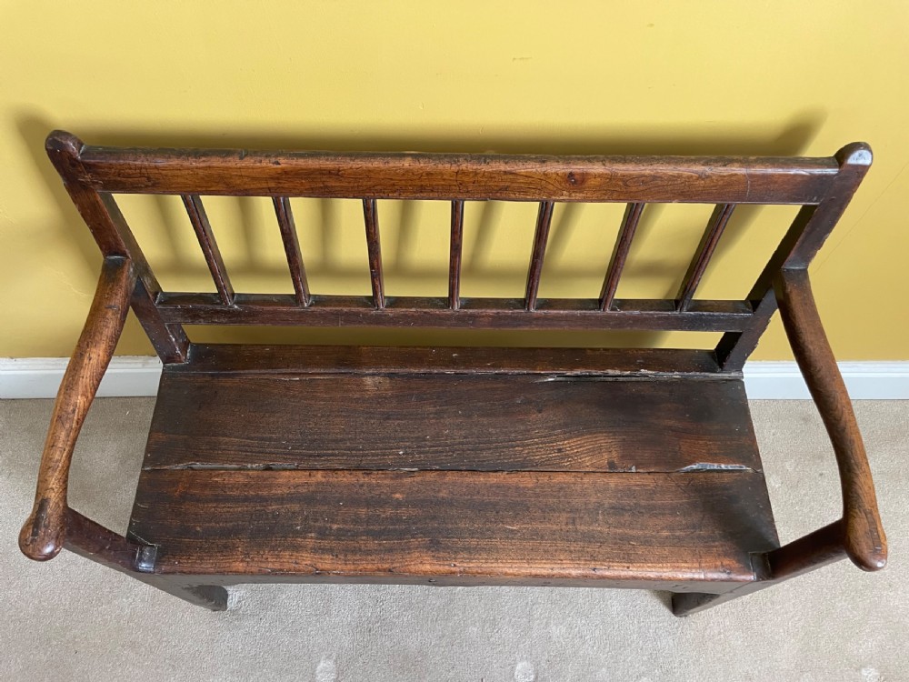 A dark oak Queen Anne period bench featuring open armrests, a vertical spindle backrest, a wooden plank seat, and a shaped apron. View IV