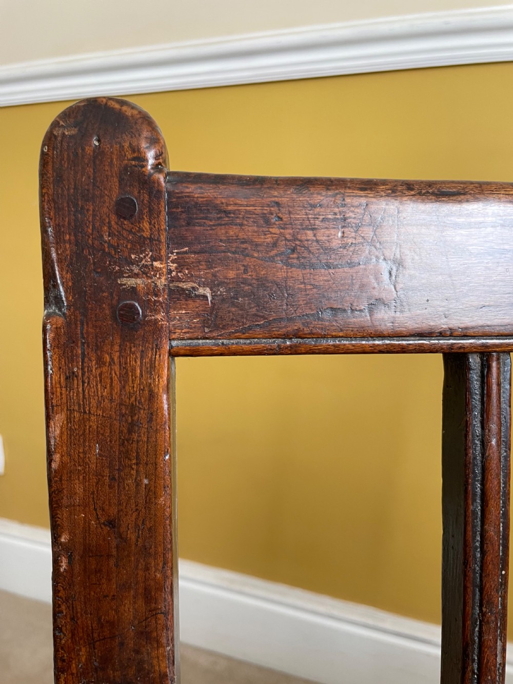 A dark oak Queen Anne period bench featuring open armrests, a vertical spindle backrest, a wooden plank seat, and a shaped apron. View VI