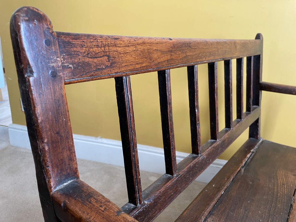 A dark oak Queen Anne period bench featuring open armrests, a vertical spindle backrest, a wooden plank seat, and a shaped apron. View VIII