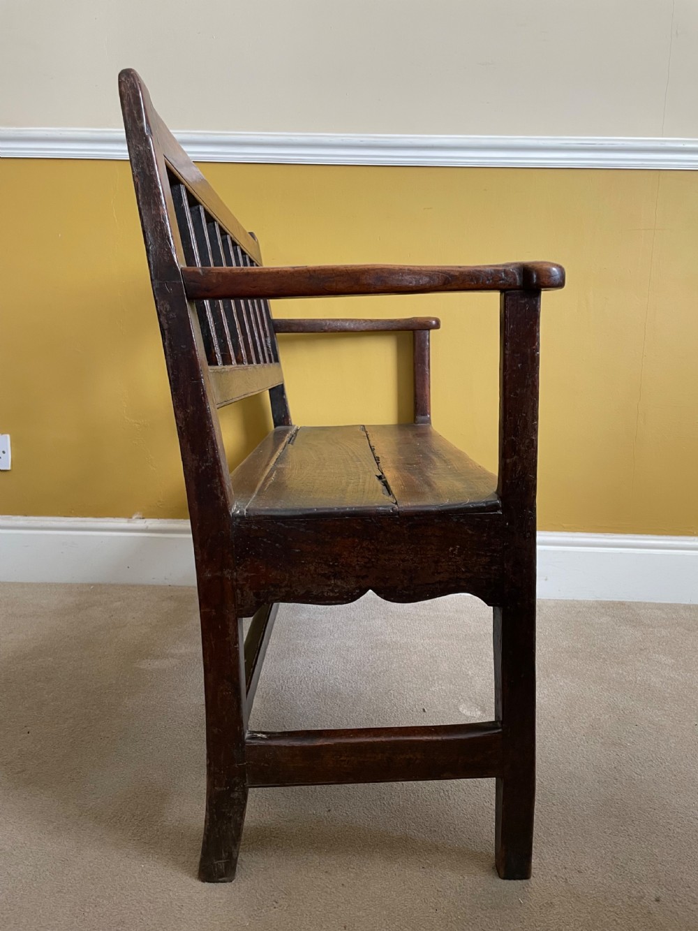 A dark oak Queen Anne period bench featuring open armrests, a vertical spindle backrest, a wooden plank seat, and a shaped apron. View IX