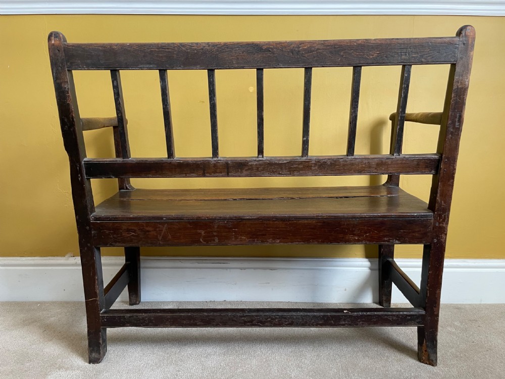 A dark oak Queen Anne period bench featuring open armrests, a vertical spindle backrest, a wooden plank seat, and a shaped apron. View X