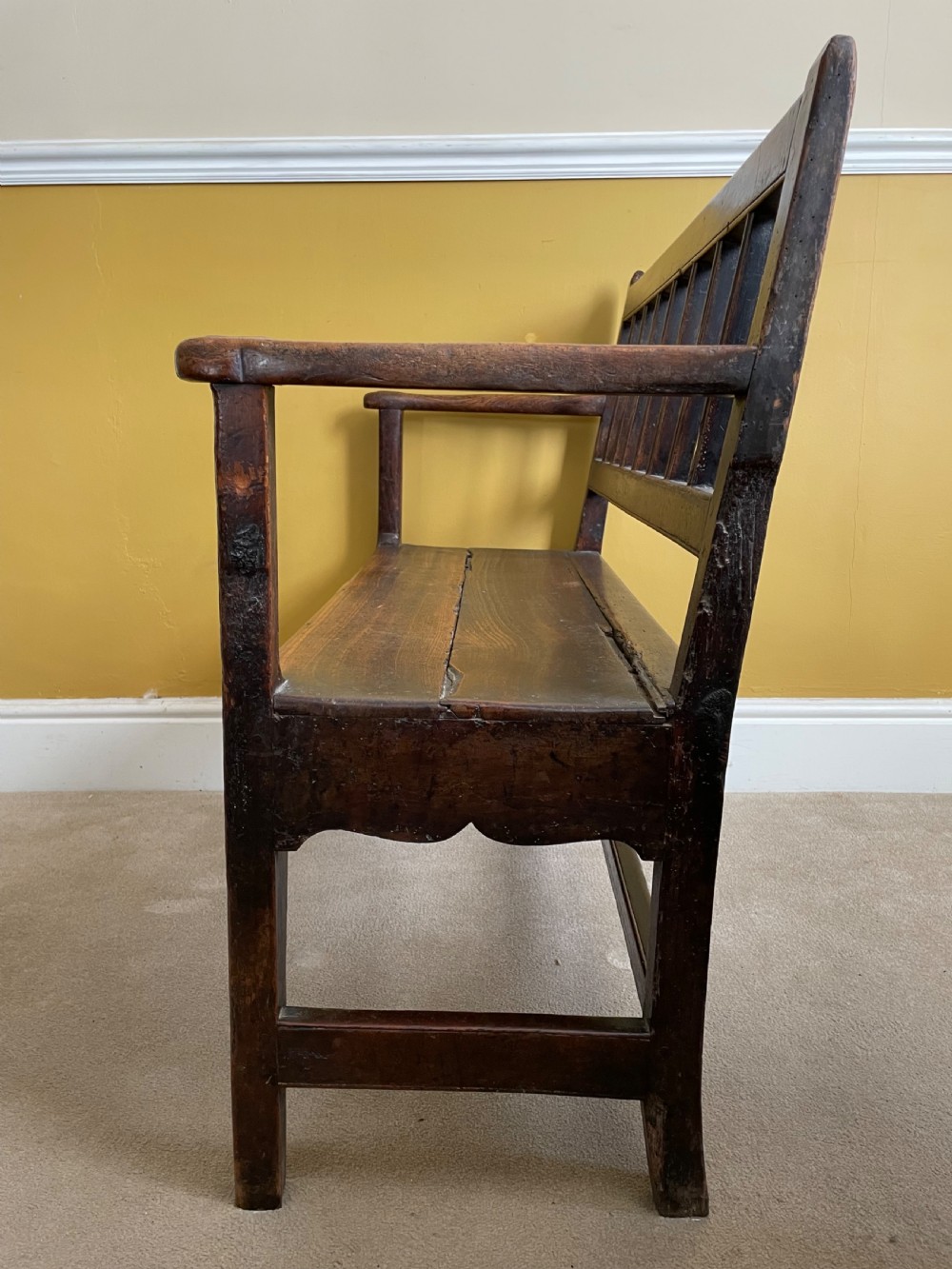 A dark oak Queen Anne period bench featuring open armrests, a vertical spindle backrest, a wooden plank seat, and a shaped apron. View XI