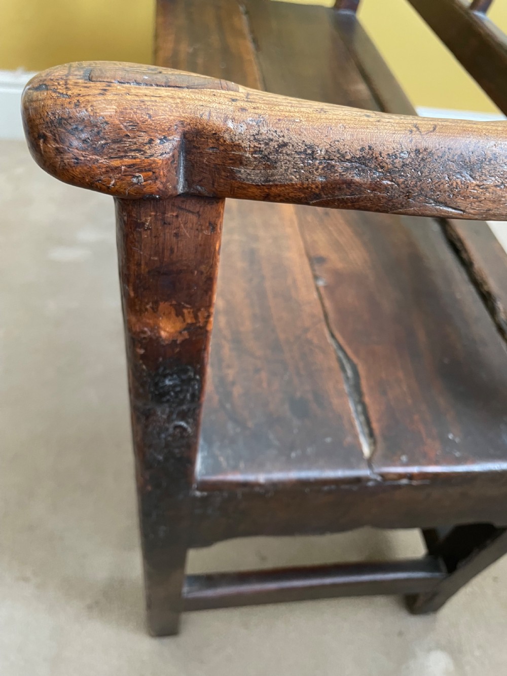 A dark oak Queen Anne period bench featuring open armrests, a vertical spindle backrest, a wooden plank seat, and a shaped apron. View XII