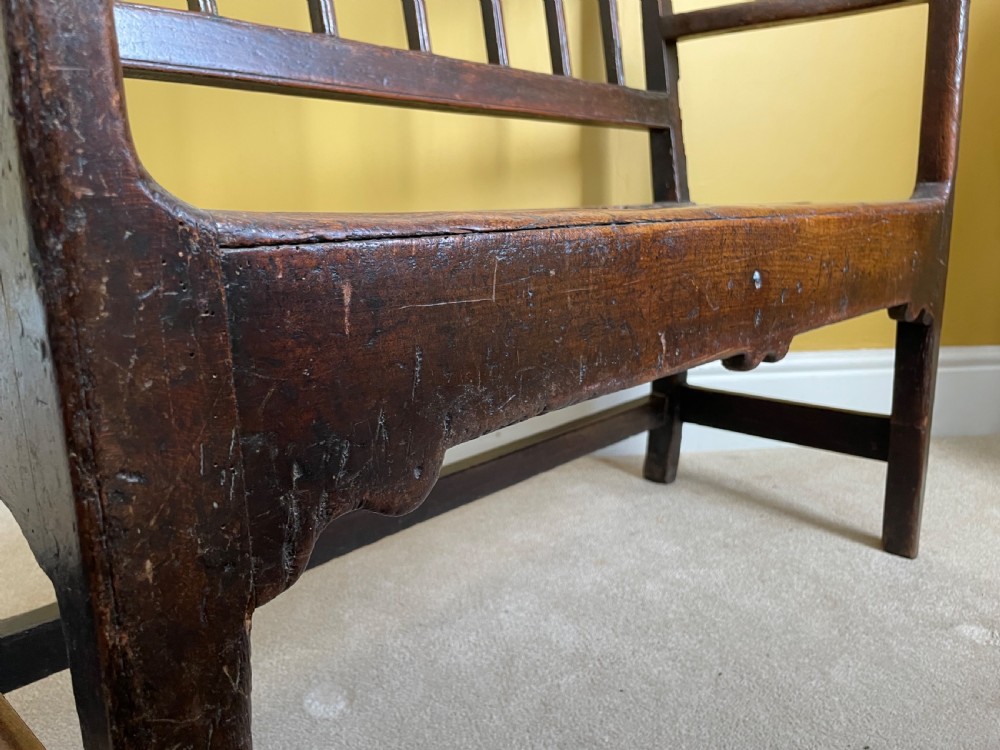 A dark oak Queen Anne period bench featuring open armrests, a vertical spindle backrest, a wooden plank seat, and a shaped apron. View XIV