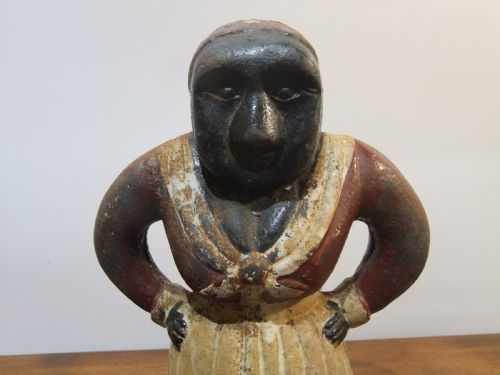 fig 1. A nineteenth-century painted cast iron money bank depicting a standing figure with hands on hips wearing a red top and cream apron.