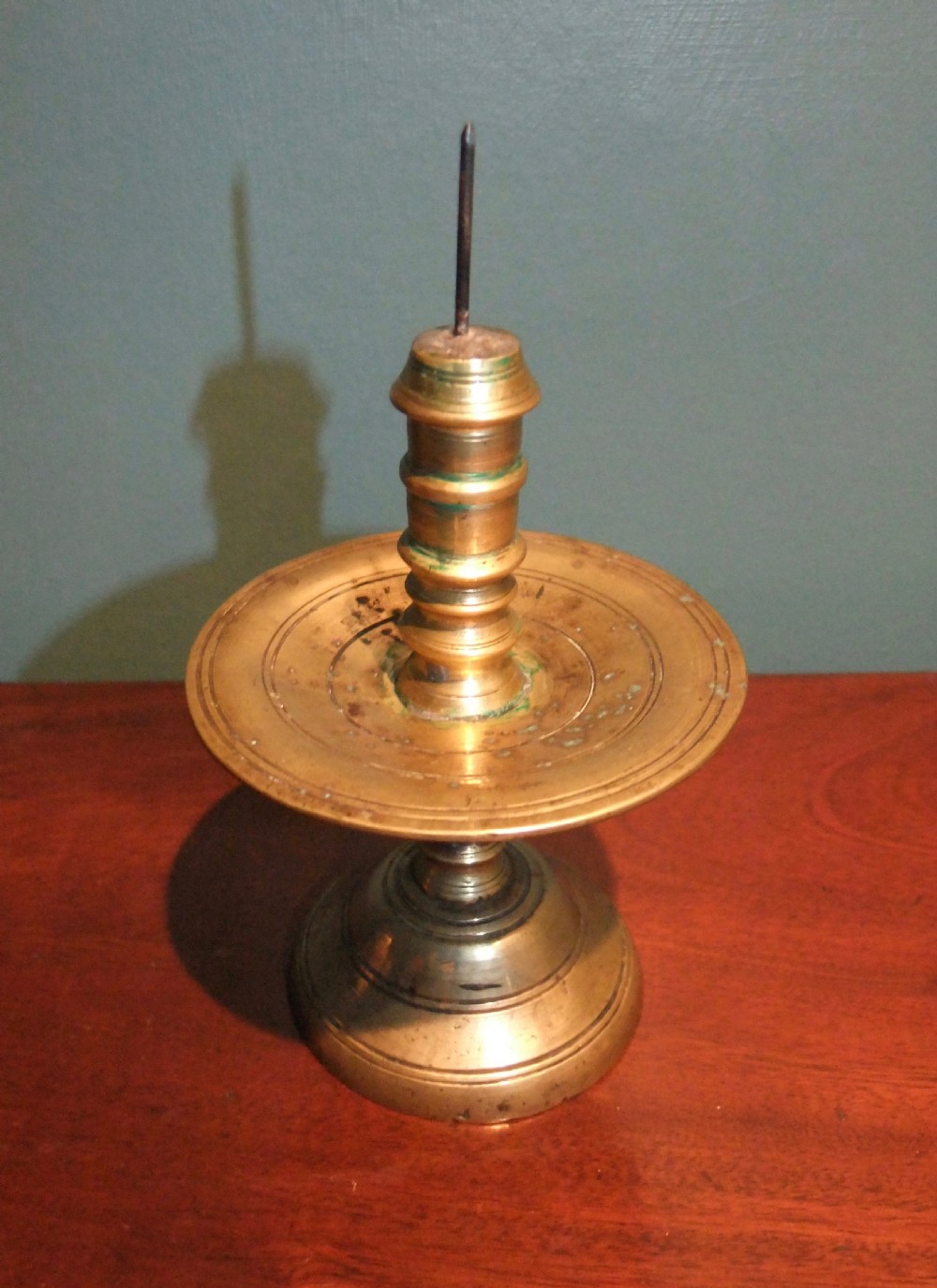 A 17th-century cast brass pricket candlestick featuring a central iron spike, a turned column stem, a wide circular drip tray, and a stepped circular base.